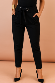 High-waisted black paperbag pants with front pleats, fabric belt with metal grommets, and a tapered ankle fit, styled with black pointed heels.