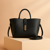 Black pebbled leather handbag with short handle, gold-tone hardware, and a central strap, styled on a display pedestal with a warm gradient backdrop.