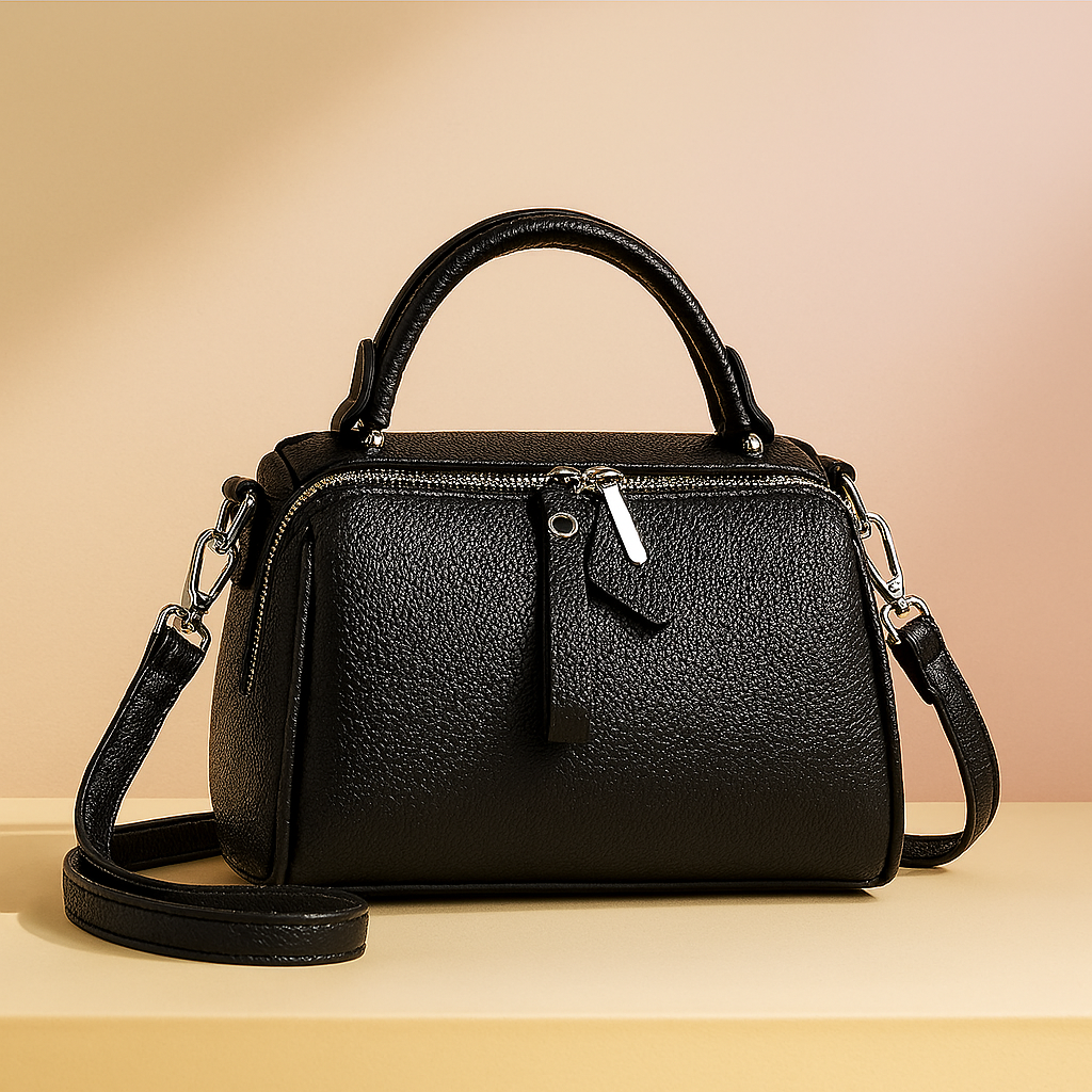 Black pebbled leather handbag with top handle, silver zipper hardware, and detachable shoulder strap, displayed on a neutral platform with a warm sand and pastel pink gradient background.