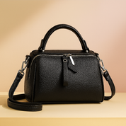 Black pebbled leather handbag with top handle, silver zipper hardware, and detachable shoulder strap, displayed on a neutral platform with a warm sand and pastel pink gradient background.