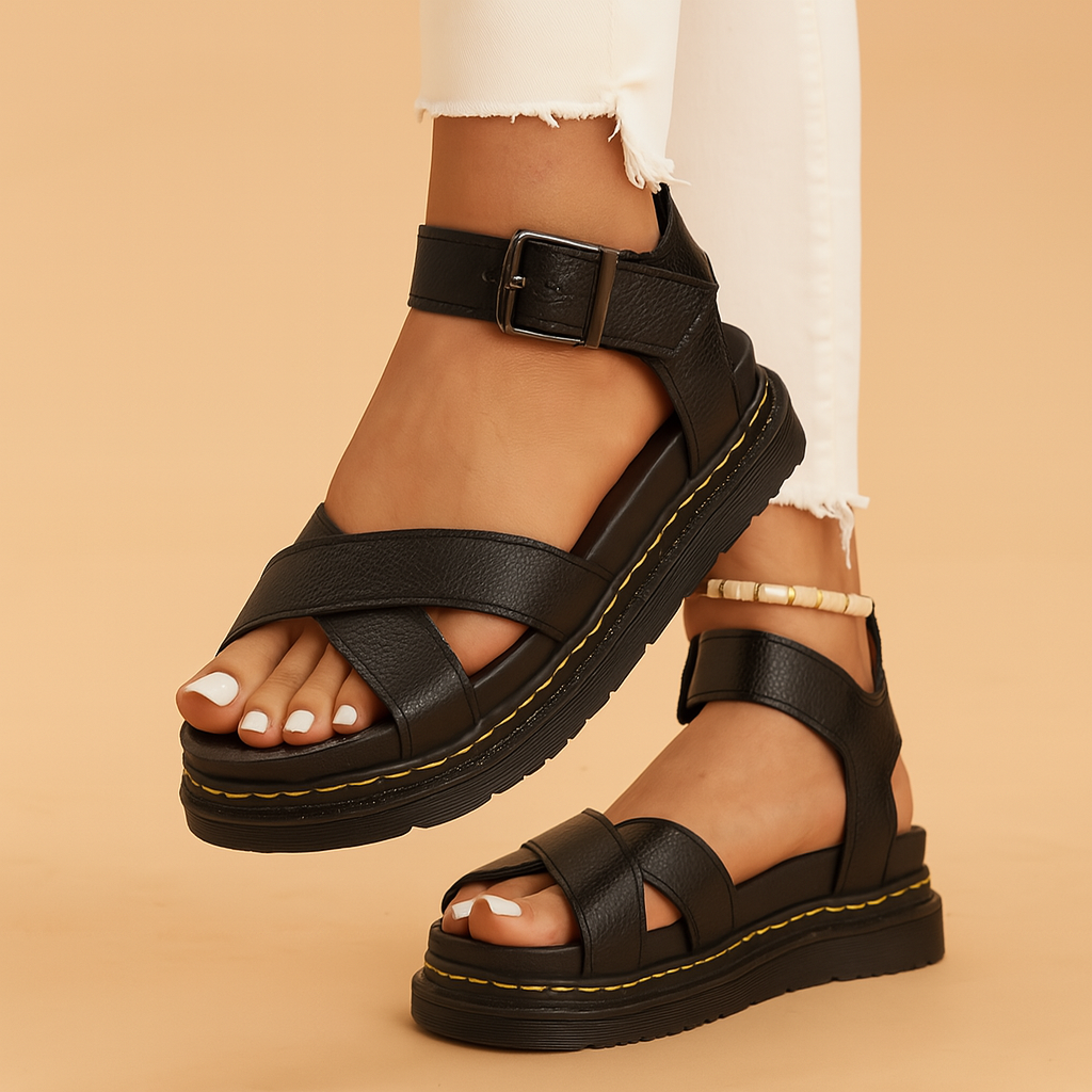 Black leather platform sandals with thick crisscross straps, ankle buckle closure, and yellow-stitched rugged soles, styled with frayed white skinny jeans and white pedicure.