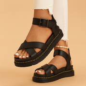 Black leather platform sandals with thick crisscross straps, ankle buckle closure, and yellow-stitched rugged soles, styled with frayed white skinny jeans and white pedicure.