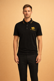 Black short-sleeve polo shirt with structured collar, contrast gold piping along the collar edge, embroidered gold crest on the chest, tonal button placket, and smooth solid fabric finish.
