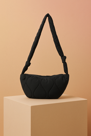 Black quilted shoulder bag with a crescent silhouette and knotted padded strap, presented on a beige platform against a warm sand and pastel pink gradient background.