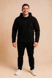 Black softshell tactical jacket with multiple zippered pockets and hood, styled with black trousers and white sneakers.