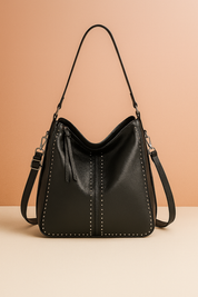 Black slouchy leather shoulder bag with silver stud trim, center seam, zipper tassel detail, and dual straps, styled on a soft gradient studio backdrop.