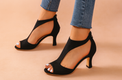 Black suede open-toe T-strap heels featuring a soft matte finish, elegant curved cutouts, and a back zipper closure, styled with a tapered mid-height heel and smooth beige outsole.