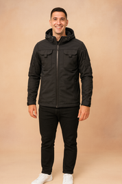 Black tactical softshell jacket with hood, chest flap pockets, and multiple zippered compartments, styled with black trousers and white sneakers.