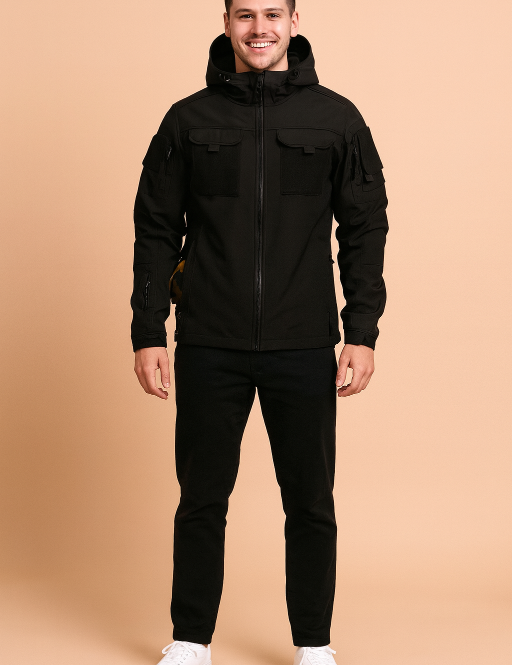 Black tactical softshell jacket with hood, Velcro chest patches, multiple zippered pockets, styled with black trousers and white sneakers.