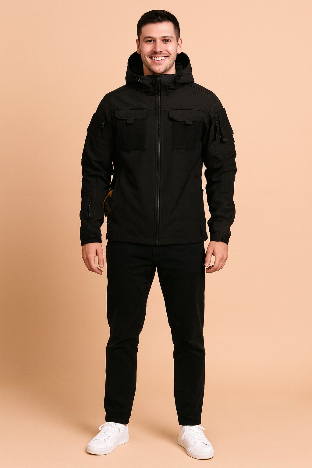 Black tactical softshell jacket with hood, Velcro chest patches, multiple zippered pockets, styled with black trousers and white sneakers.