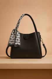 Black textured shoulder bag with a curved top handle, side strap, and monochrome diamond-patterned scarf tied to the handle, displayed on a neutral platform.