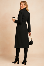 Black longline trench coat with a waist belt, paired with black fitted pants and black suede block-heel ankle boots, accessorized with a black crocodile-embossed handbag with gold chain strap.