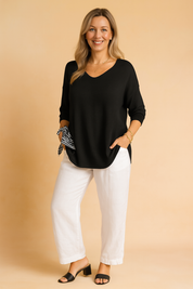 Black V-neck long-sleeve blouse with relaxed fit, paired with white straight-leg linen trousers and black heeled sandals, accented by a striped scarf tied at the wrist.