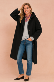 Black wool button-up coat with a relaxed oversized fit, wide sleeves, side pockets, and front button closure, styled over a light gray V-neck sweater, paired with medium-wash straight-leg jeans, a colorful paisley scarf, and black leather flats.