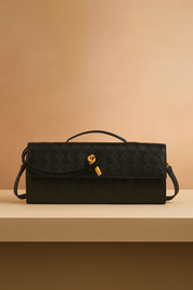 Rectangular black woven leather handbag with gold knot detail and flap closure, displayed on a beige platform against a pastel sand-to-pink gradient background.