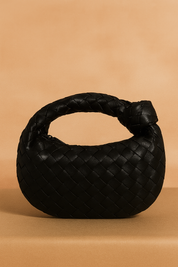 Black woven leather hobo handbag with rounded structure and knotted handle, styled on a warm pastel tabletop.