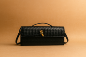 Rectangular black woven leather handbag with top handle, gold-tone hardware, and wraparound closure detail, displayed on a warm sand and pastel peach gradient background.
