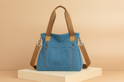 Blue canvas tote bag with brown handles and silver zippers, set on a cream platform against a soft beige gradient background.