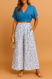 Two-piece summer set featuring a cropped button-up blue blouse with cuffed short sleeves and high-waisted wide-leg trousers in a white base with soft blue botanical print.