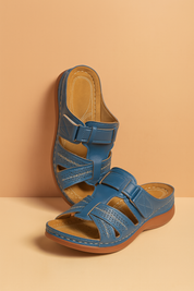 Blue leather open-toe slide sandals with crisscross perforated straps, a secure hook-and-loop closure, cushioned tan insole with visible stitching, and brown low wedge outsole.