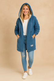 Cozy oversized blue sherpa hoodie jacket with long sleeves, ribbed cuffs, kangaroo-style pockets, and embroidered back detail. Styled with fitted blue jeans and white sneakers for a casual winter look.