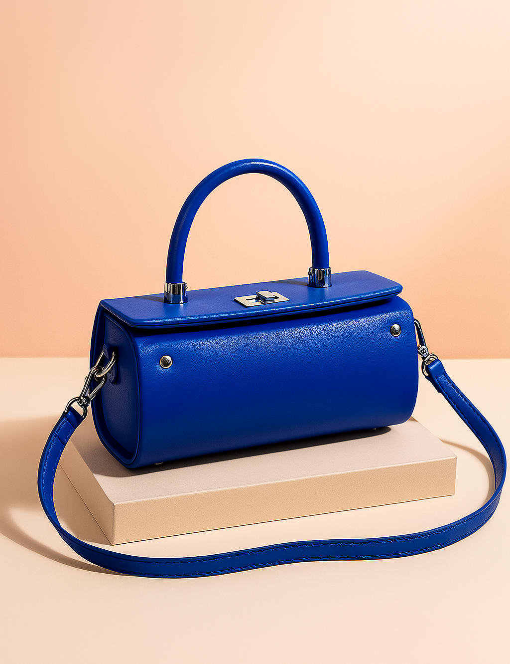 Glossy blue structured handbag with a cylindrical body, top handle, silver-tone hardware, and detachable strap, styled on a neutral backdrop for a premium product showcase.