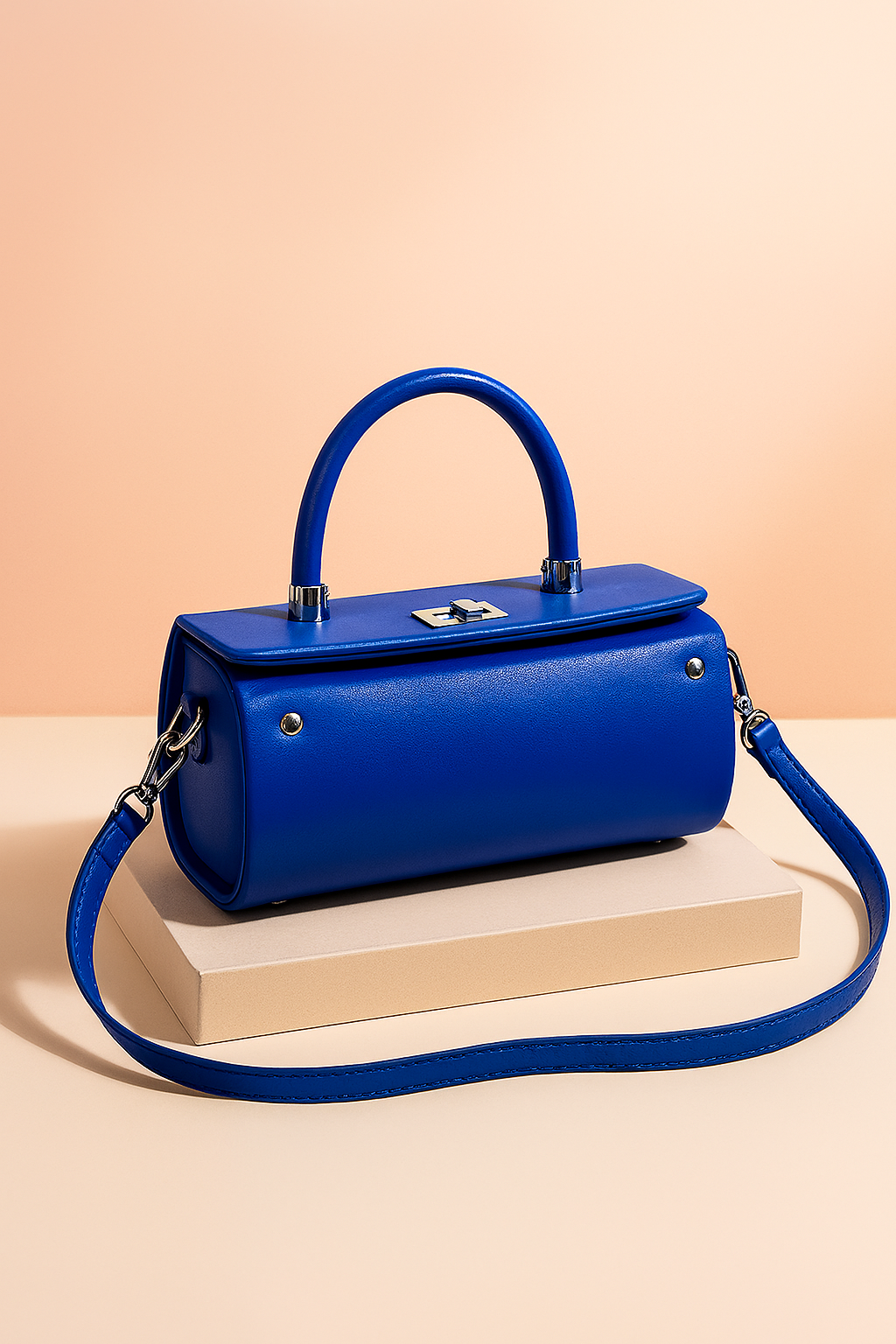 Glossy blue structured handbag with a cylindrical body, top handle, silver-tone hardware, and detachable strap, styled on a neutral backdrop for a premium product showcase.