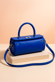 Glossy blue structured handbag with a cylindrical body, top handle, silver-tone hardware, and detachable strap, styled on a neutral backdrop for a premium product showcase.