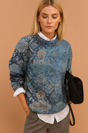 Blue vintage-patterned crewneck sweater with intricate mandala and floral motifs, layered over a crisp white collared shirt and paired with tailored grey trousers.