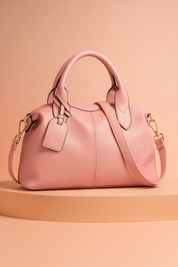 Blush pink leather handbag with top handles, gold hardware, and detachable shoulder strap, styled on a pastel platform with warm gradient lighting.