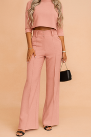 High-waisted, wide-leg tailored pants in blush pink with belt loops, center seam detailing, and a clean front closure, styled with black open-toe heels and a minimalist handbag.