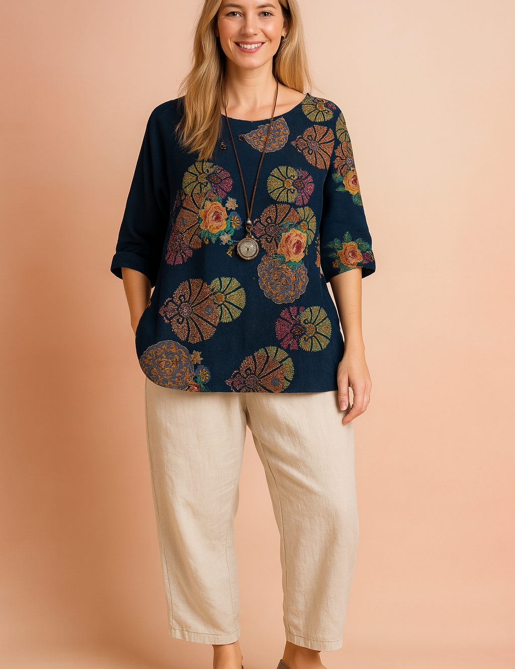 Navy blue asymmetrical blouse with rolled-up sleeves, detailed with a vibrant mandala and floral print featuring roses and fan motifs in red, green, yellow, and violet. Decorated with small buttons along the neckline and styled with a long pendant necklace, the top is paired with loose cream-colored trousers.