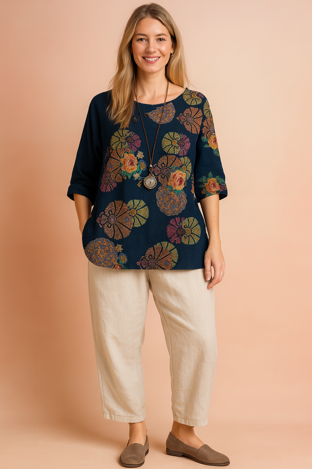 Navy blue asymmetrical blouse with rolled-up sleeves, detailed with a vibrant mandala and floral print featuring roses and fan motifs in red, green, yellow, and violet. Decorated with small buttons along the neckline and styled with a long pendant necklace, the top is paired with loose cream-colored trousers.