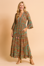 Bohemian-style maxi dress with flared sleeves, gathered waistline, and vibrant paisley pattern in earthy red, turquoise, and green tones, featuring front tassel ties and a tiered hem.