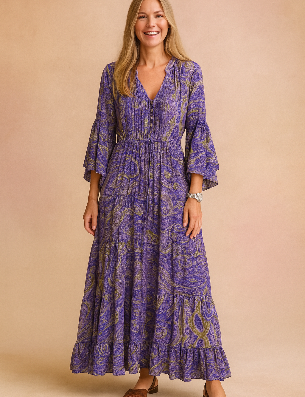 Bohemian-inspired long maxi dress with tiered hem, featuring a paisley print in purple, blue, and olive green tones, long flared sleeves, front buttons, and an adjustable drawstring waist.