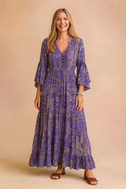 Bohemian-inspired long maxi dress with tiered hem, featuring a paisley print in purple, blue, and olive green tones, long flared sleeves, front buttons, and an adjustable drawstring waist.