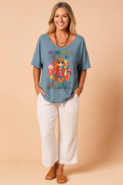 Loose-fit blue graphic T-shirt featuring a colorful boho print and bold lettering, paired with relaxed white linen pants and styled for casual summer wear.