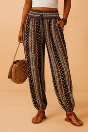 Bohemian high-waist drawstring jogger pants featuring vertical tribal-inspired patterns in black, cream, and rust tones with a shirred waistband and three buttons, styled with brown flat sandals.