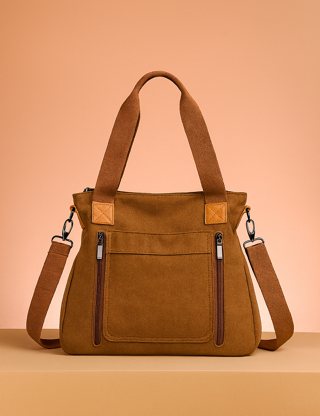 Brown canvas tote bag with twin zippered front pockets, canvas handles, and adjustable crossbody strap on a warm pastel tabletop.