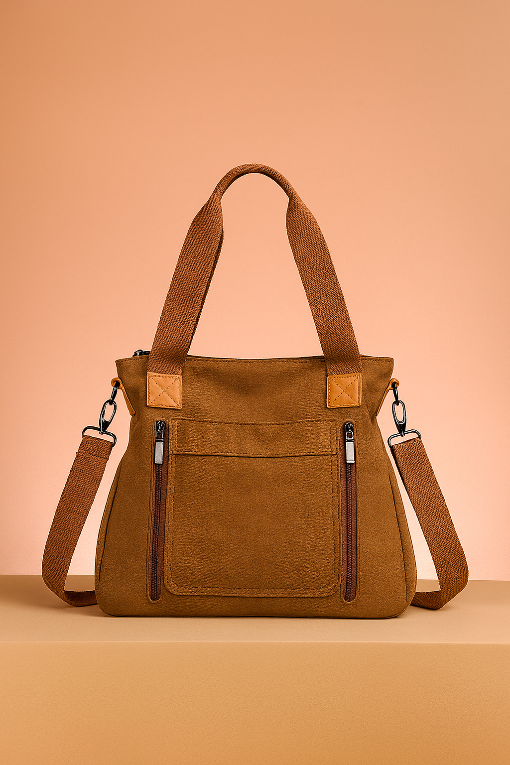 Brown canvas tote bag with twin zippered front pockets, canvas handles, and adjustable crossbody strap on a warm pastel tabletop.