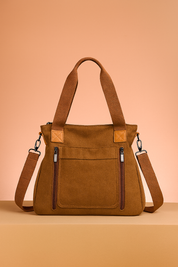 Brown canvas tote bag with twin zippered front pockets, canvas handles, and adjustable crossbody strap on a warm pastel tabletop.