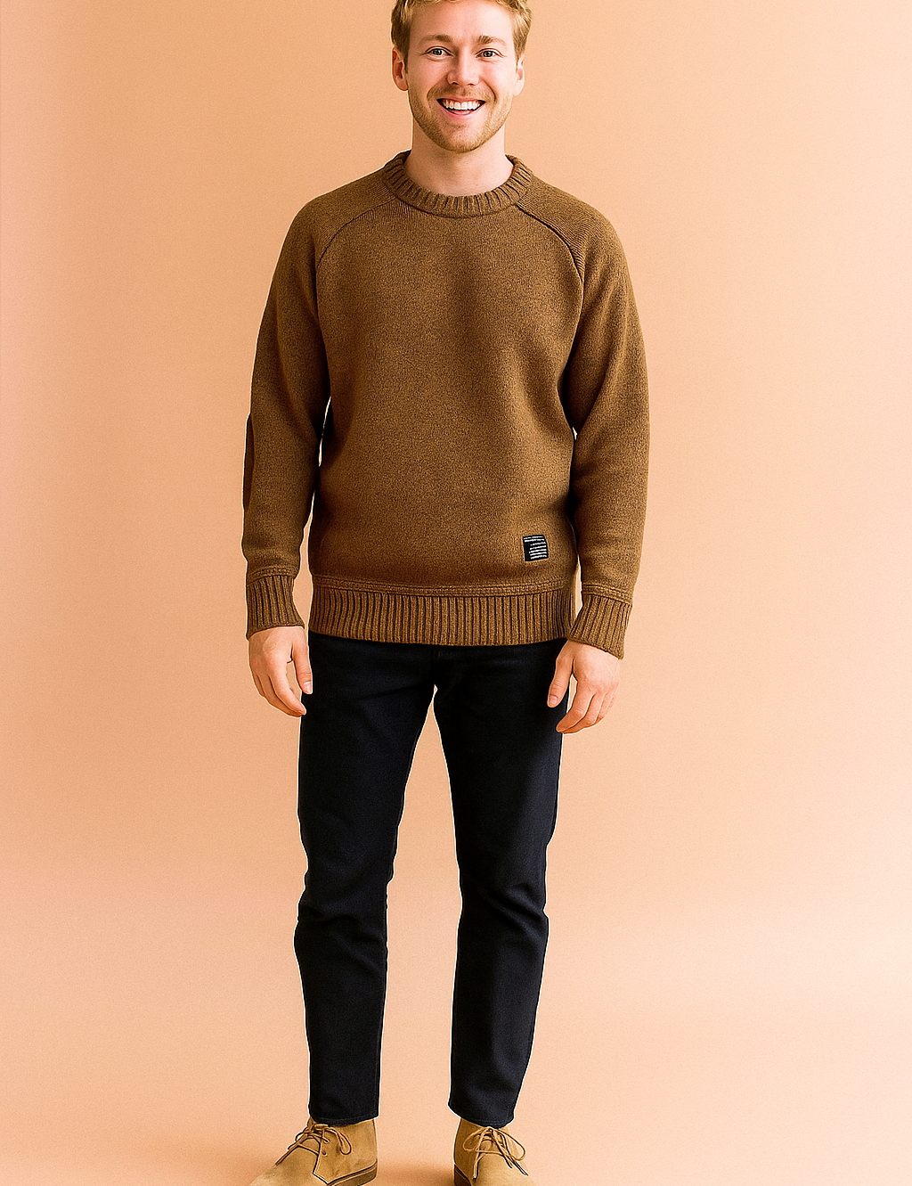 Brown crewneck sweater with ribbed cuffs and hem, suede elbow patches, and a small black label on the lower front, paired with dark slim-fit trousers and beige suede desert boots