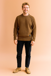 Brown crewneck sweater with ribbed cuffs and hem, suede elbow patches, and a small black label on the lower front, paired with dark slim-fit trousers and beige suede desert boots
