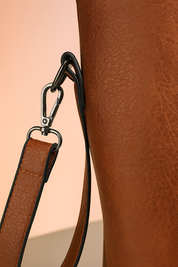 Close-up of a brown leather bag’s strap attachment showing a silver clasp and black metal loop, against a warm sand and pastel pink gradient background.