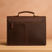 Brown leather briefcase with zipper pocket and top handle, posed on a wood surface against a softly lit warm sand and pastel blush gradient background.