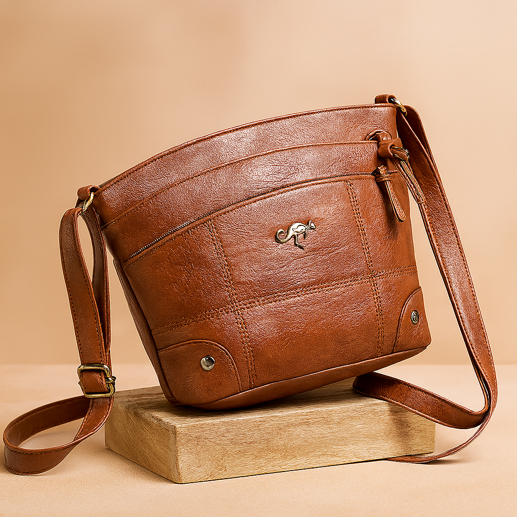 Brown leather crossbody bag with curved top zip, quilted paneling, silver kangaroo emblem, and adjustable strap, styled on a wood platform.