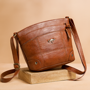 Brown leather crossbody bag with curved top zip, quilted paneling, silver kangaroo emblem, and adjustable strap, styled on a wood platform.