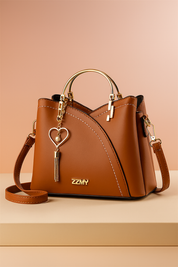 Caramel brown leather handbag with curved white stitching, gold top handles, heart-and-pearl tassel charm, and detachable strap, styled on a beige table with a soft pastel peach gradient background.