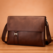 Brown leather messenger bag with silver front buckle and dark brown shoulder strap, set against a soft gradient beige and pastel pink background with warm lighting on a wood platform.