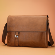 Sleek brown leather messenger bag with fold-over flap, stitched detailing, and adjustable shoulder strap, set on a soft sand-pink gradient background with elegant lighting.